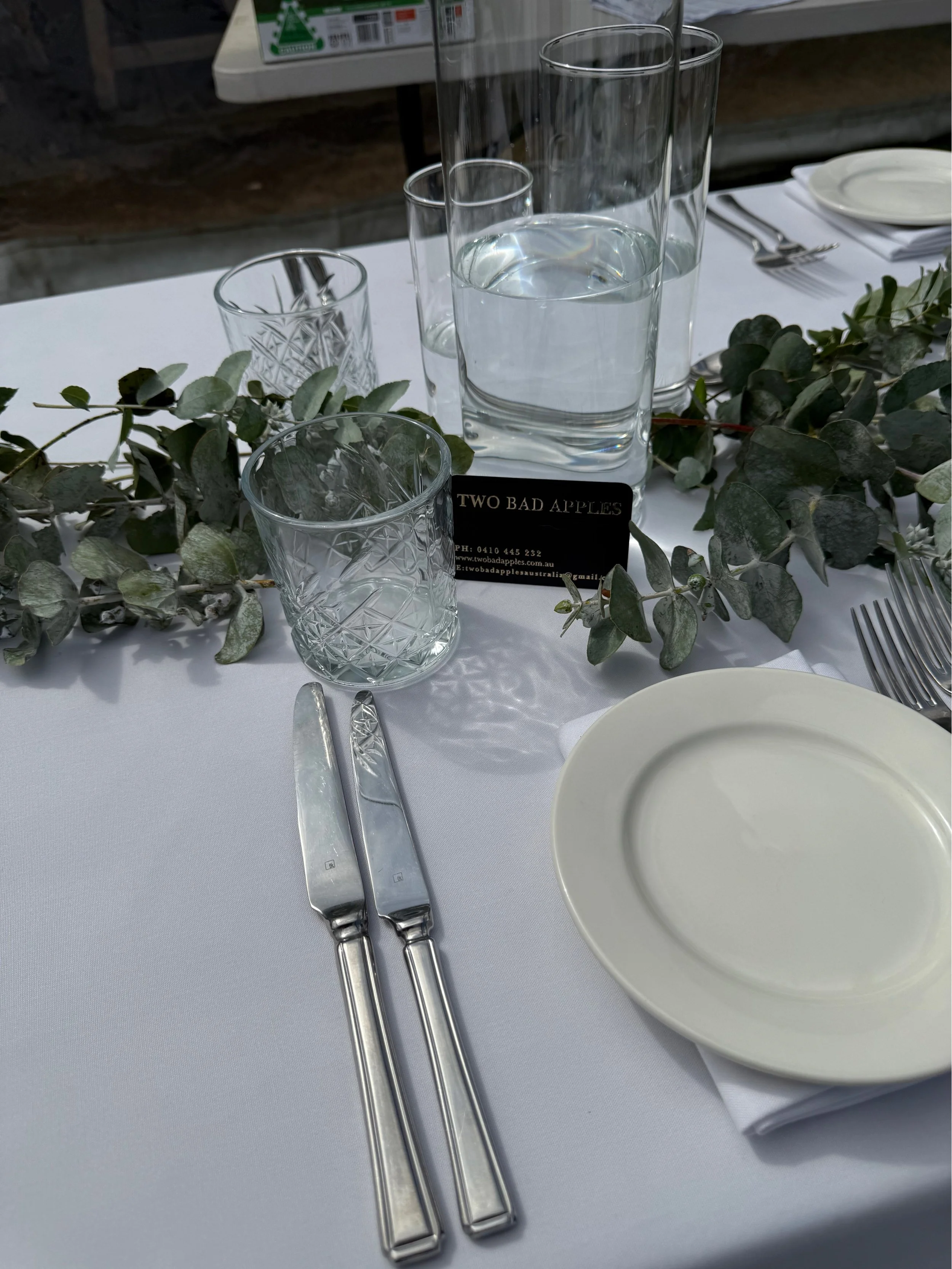 A dining table set with glassware, including five empty drinking glasses of various heights, a white plate, and silverware. Green leaves are arranged as decor around the glasses, and a business card with the text 'TWO BAD APPLES' is placed on the tab