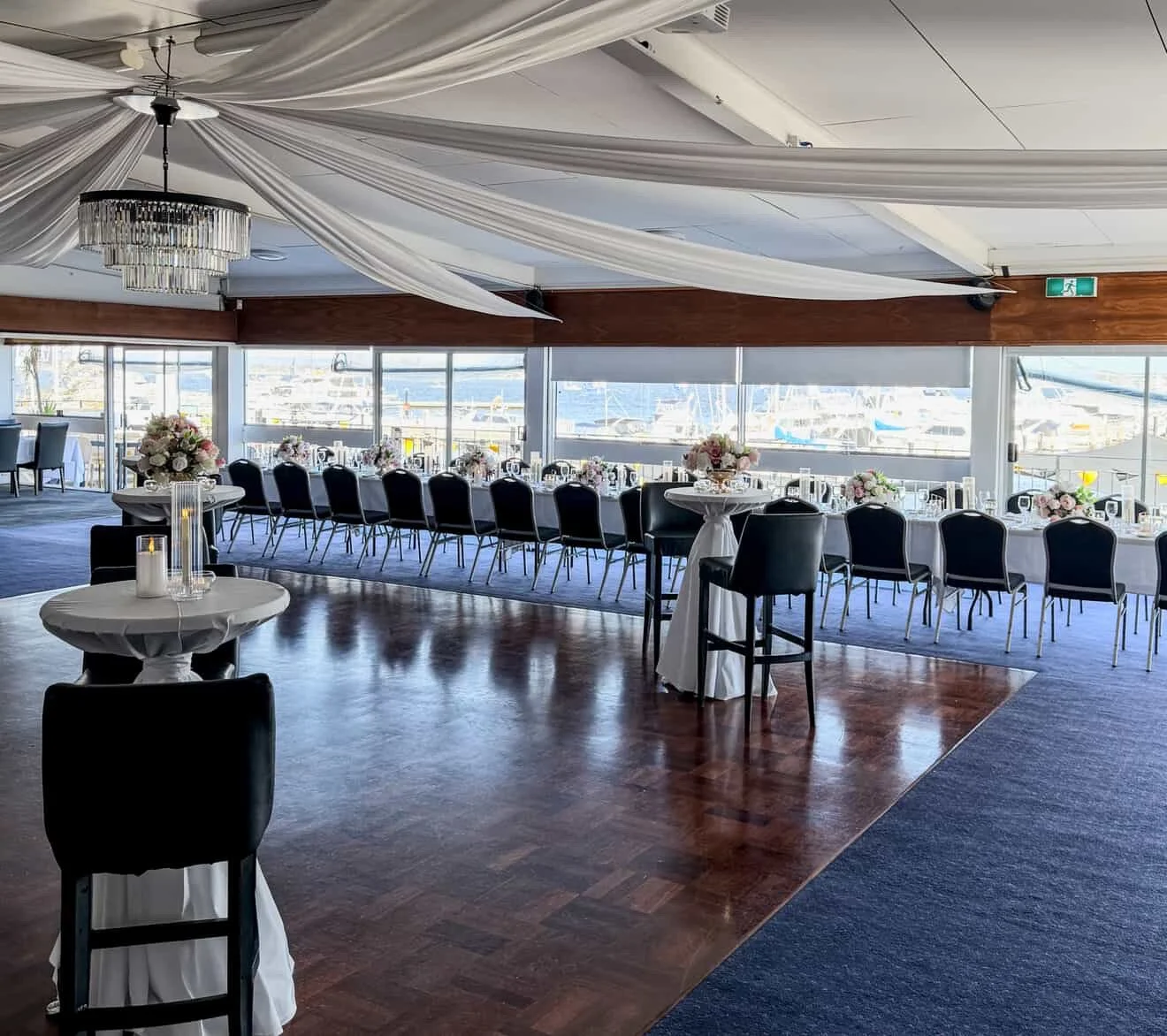 Elegant banquet hall with long tables decorated with flowers, set for a formal event, overlooking a marina through large windows.