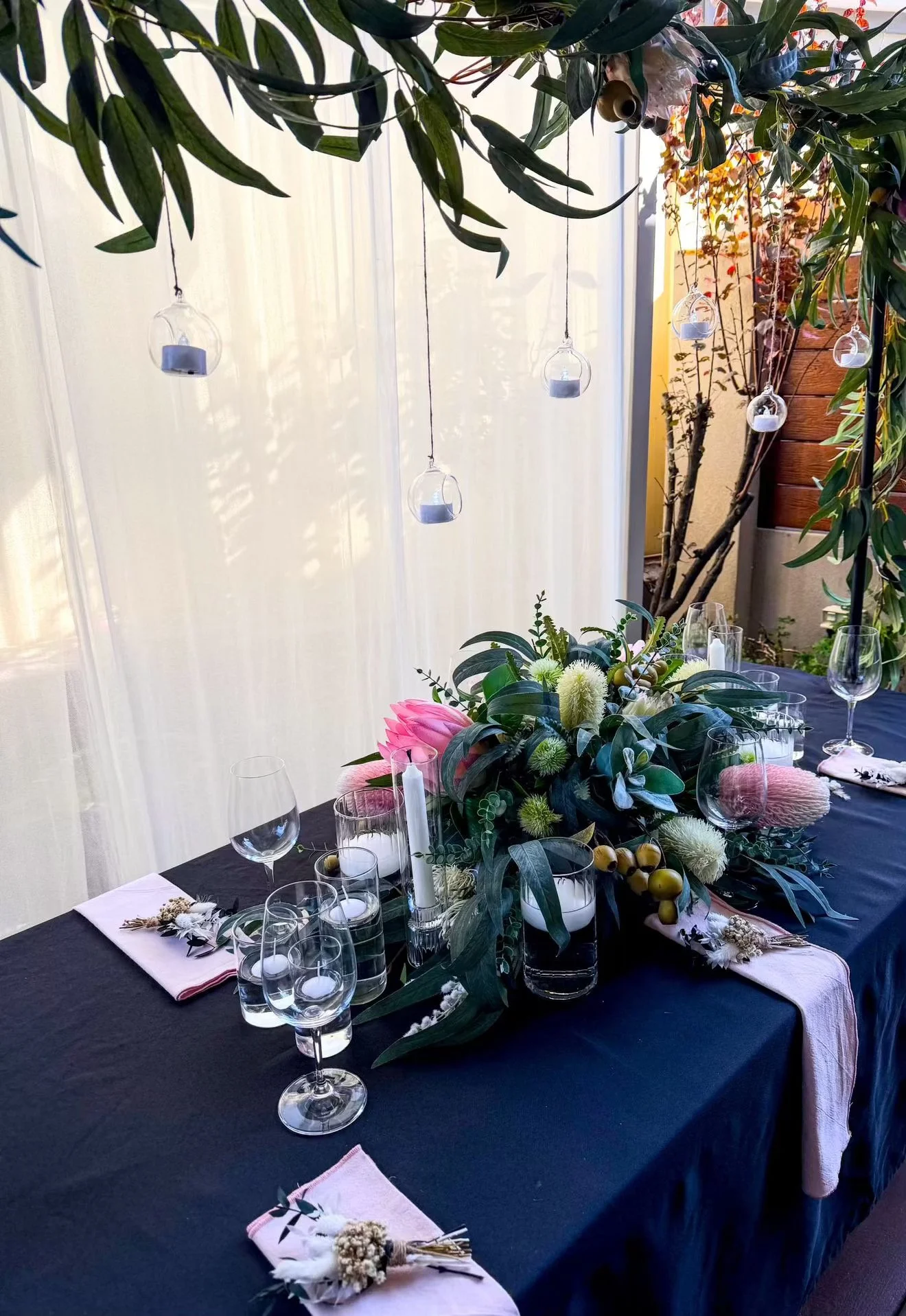 A decorated dining table set for a celebration, featuring a black tablecloth, white napkins with floral accents, multiple glassware including wine glasses, and a central floral arrangement with pink, white, and green flowers and foliage. There are sm