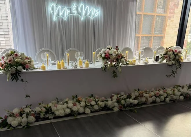 Wedding reception table with floral arrangements, candles, and a wedding neon sign in the background.