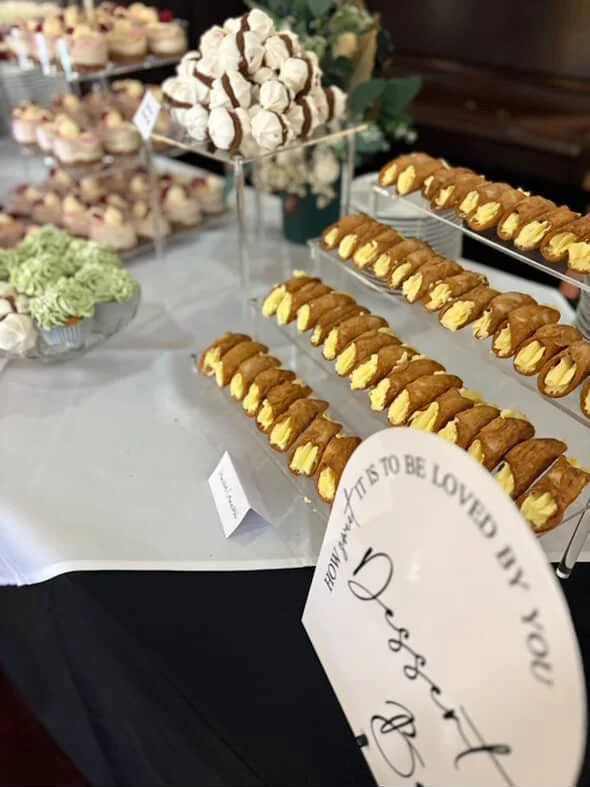 Display of assorted desserts, including cannoli and meringue cookies, arranged on tiered trays at a buffet or dessert table.