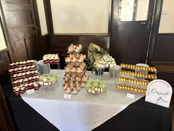 Dessert table with various cakes, cupcakes, flowers, and desserts displayed in an indoor setting.