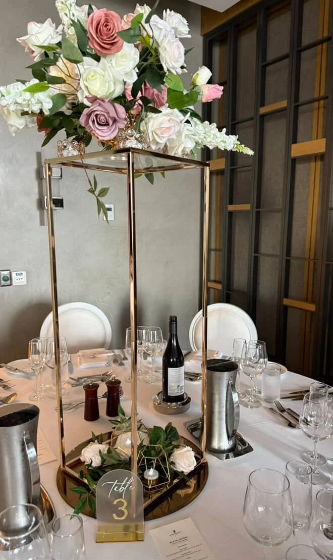 Elegant table setting with a tall floral centerpiece consisting of pink, purple, and white roses. The table has wine glasses, water glasses, silverware, a bottle of wine, salt and pepper shakers, and a table number sign indicating 'Table 3'.