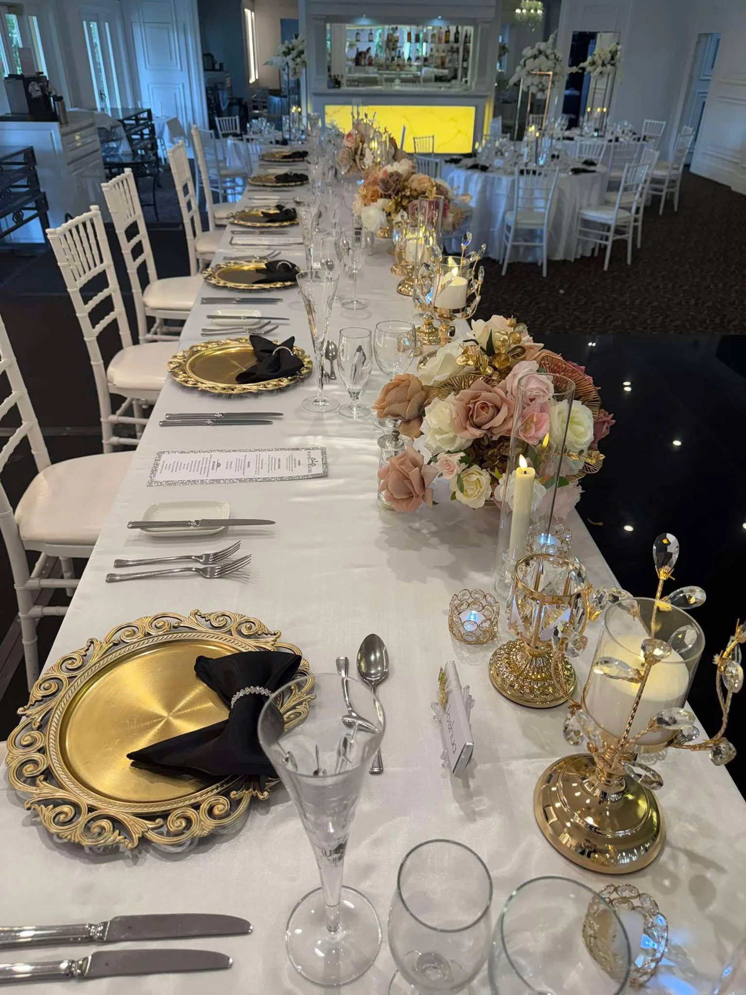 Elegant banquet table set with gold and black plates, gold candleholders, floral arrangements, and crystal glassware, in a decorated event space.