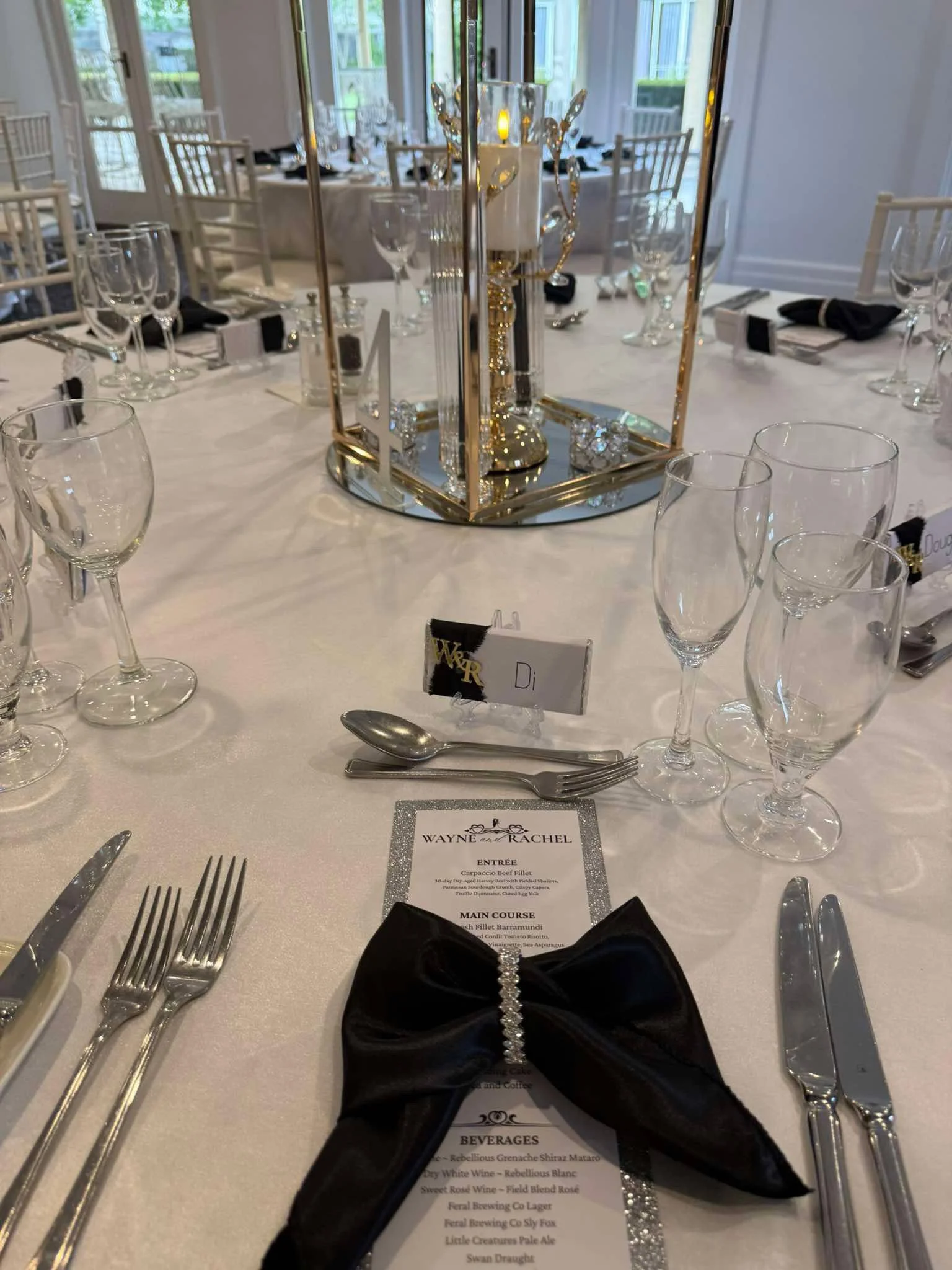 Elegant banquet table setup with glassware, cutlery, black napkins, and a centerpiece featuring a candle and decorative accents; a menu card is also visible on the table.