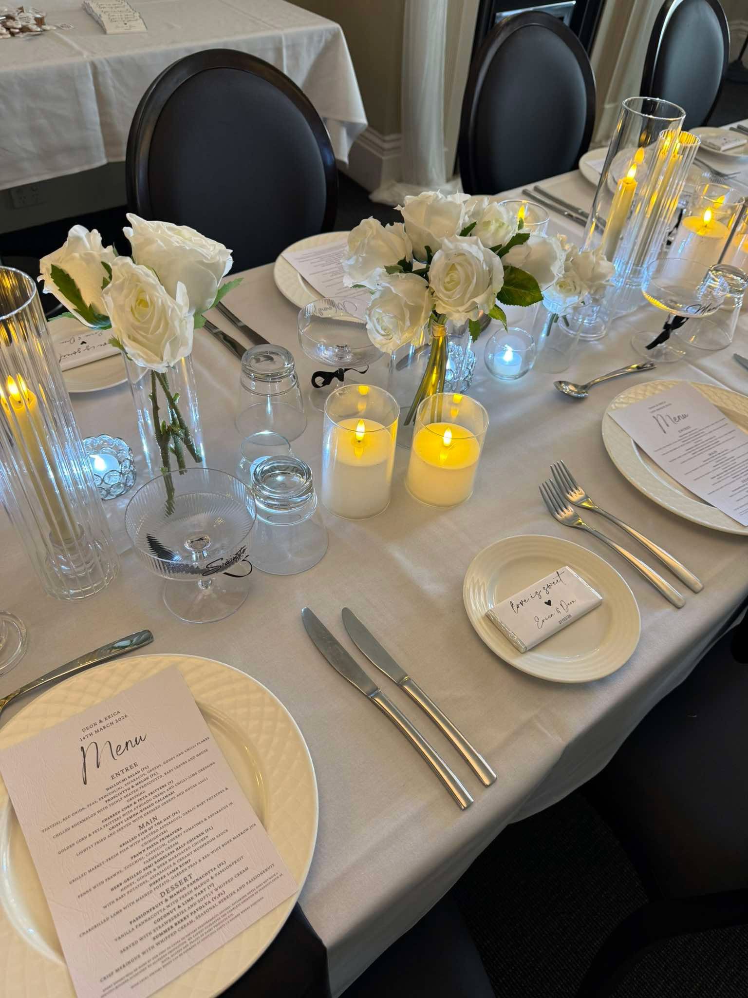A table set for a formal wedding reception with white floral centerpieces, candles, white plates with printed wedding menus, and silver utensils arranged in a formal manner.