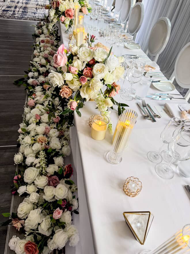Long banquet table decorated with white and pink floral arrangements, candles, and glassware for a formal event.