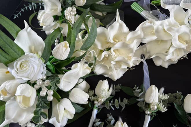 An arrangement of white flowers, including roses and calla lilies, with green foliage and pearl accents, likely for a special occasion.