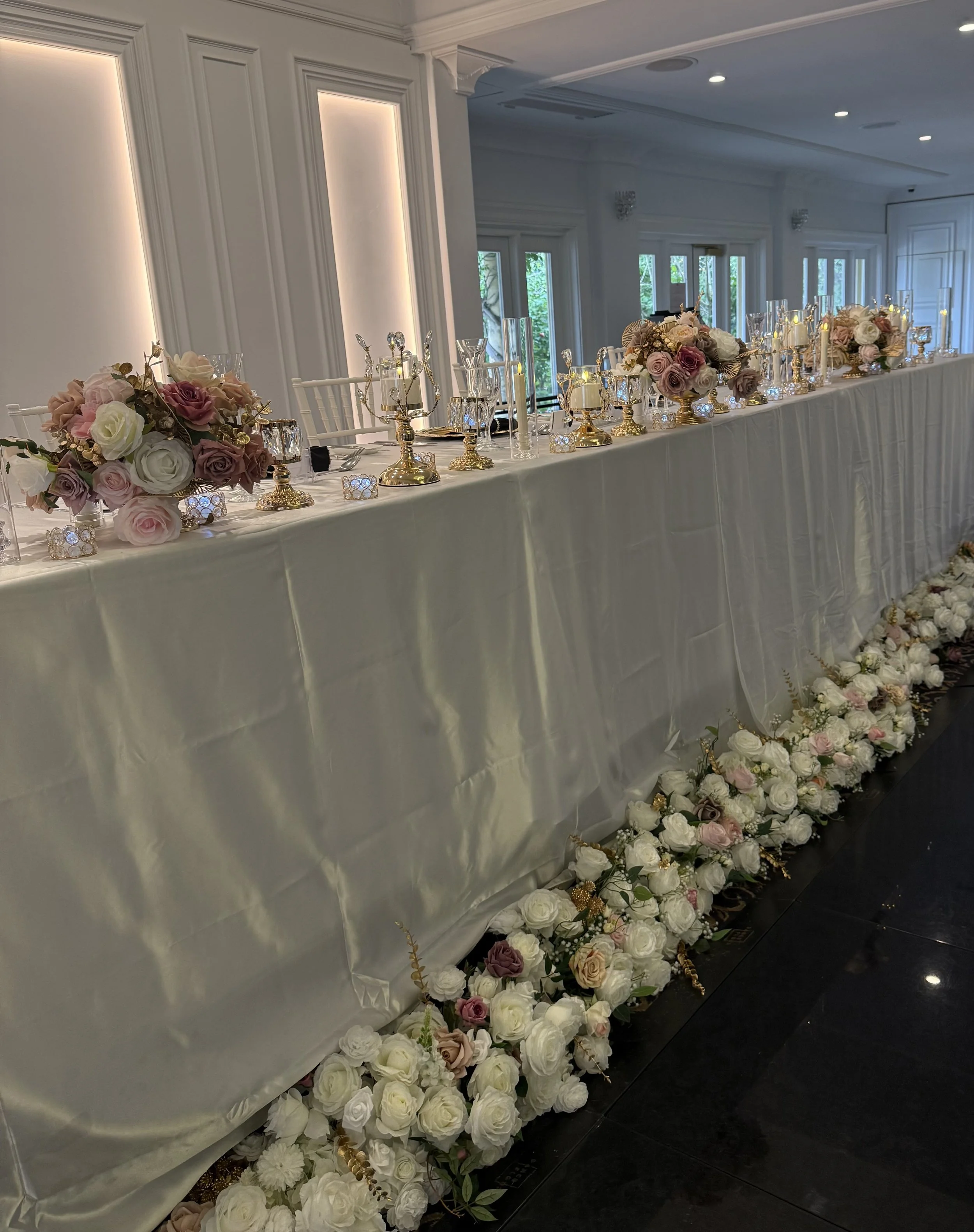 A long table decorated with floral arrangements of roses in shades of white, pink, and purple, and gold candle holders, set in an elegant, well-lit room with white walls and large windows.