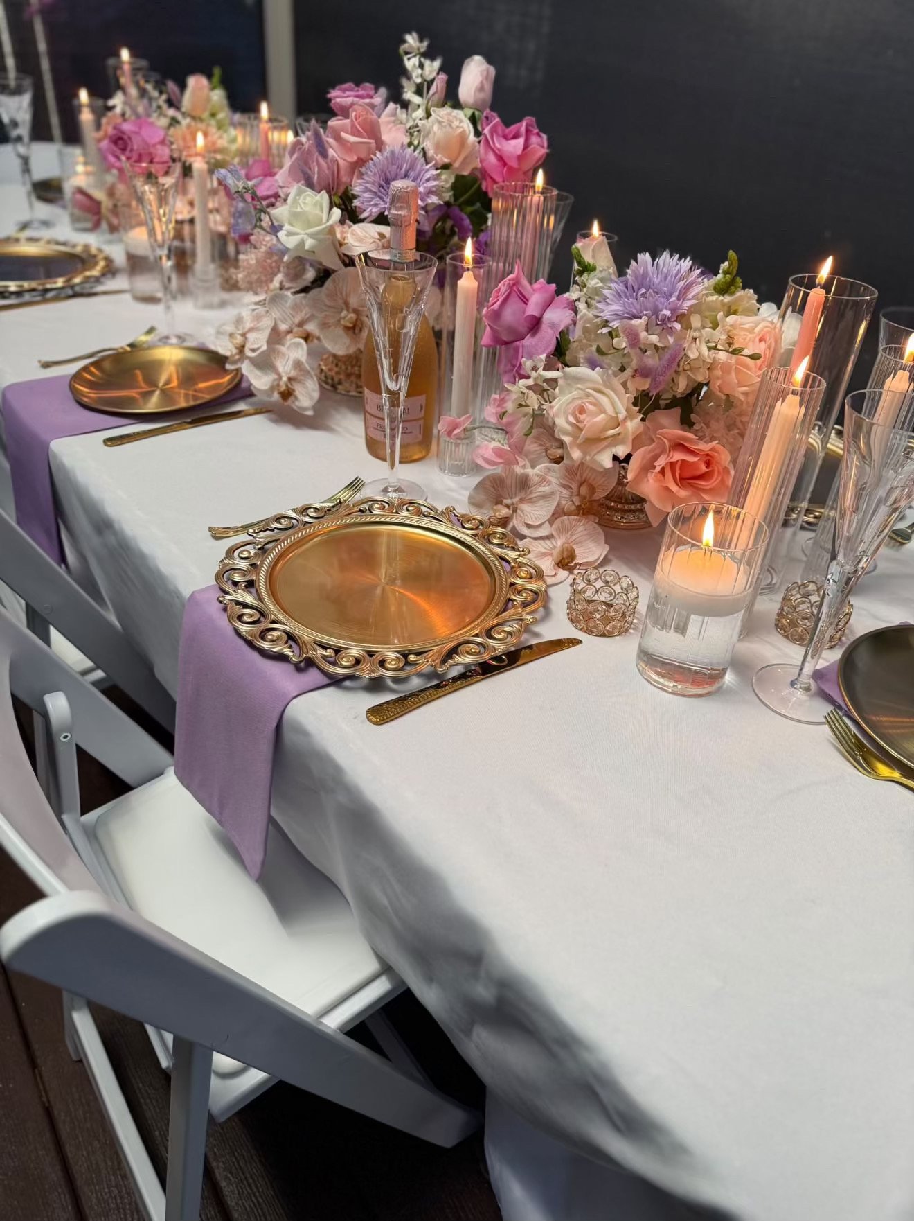 A decorated dining table with white tablecloth, lavender napkins, gold plates, gold utensils, tall candles in glass holders, and floral centerpieces with pink, purple, and white flowers.