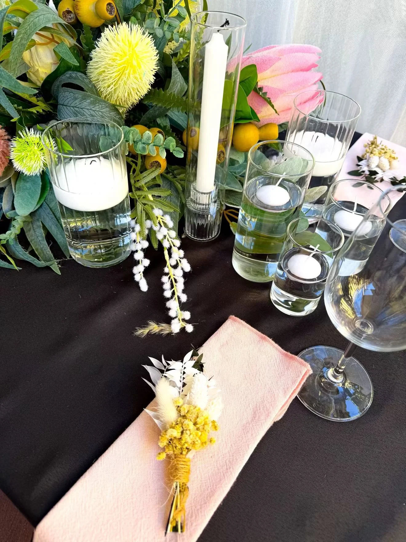 A table decorated with a floral centerpiece, white candles in glass holders, and a pink napkin with a boutonniere. The centerpiece includes various green leaves, yellow and pink flowers, and small yellow berries. The table surface is black, and there