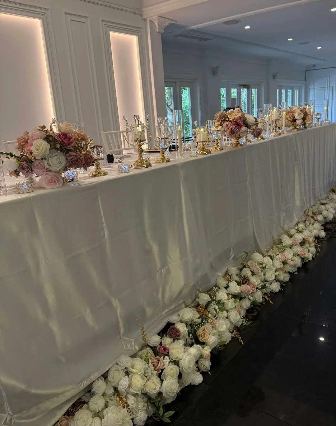 Elegant wedding reception table with white tablecloth, floral arrangements of pink and white roses, gold candle holders, and glassware, decorated with a floral garland at the base.