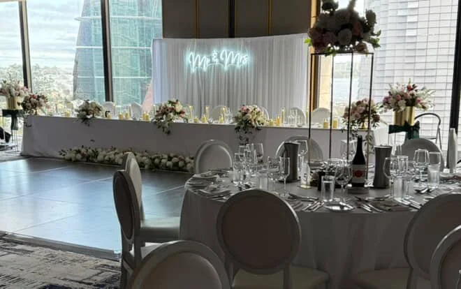 Elegant banquet table setup with floral centerpieces, glassware, and silverware, in front of a white backdrop with neon sign that says 'Mr & Mrs,' in a modern high-rise event space with city views.