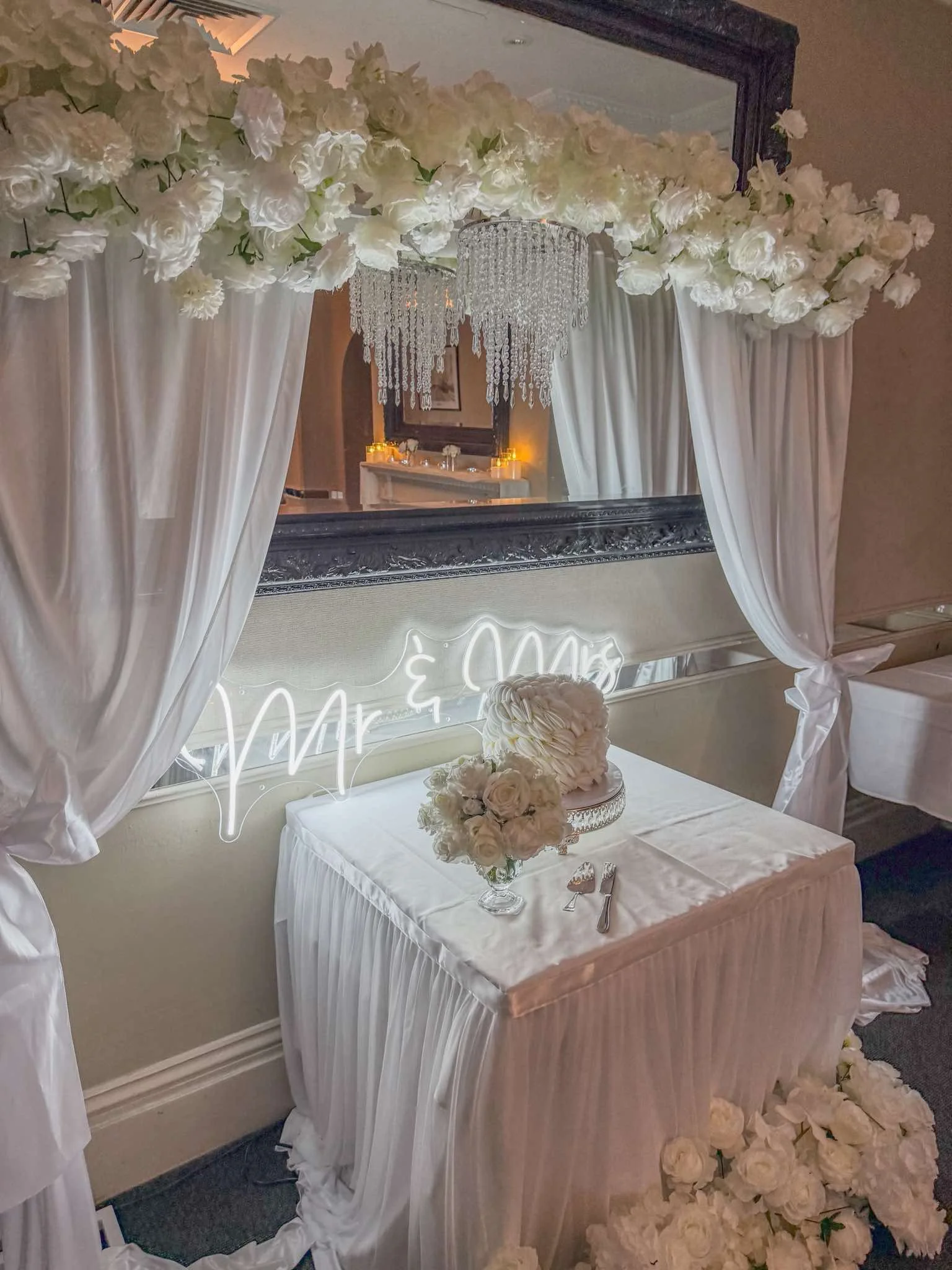A wedding head table decorated with white roses, draped white curtains, a large mirror, and a chandelier with hanging crystals.