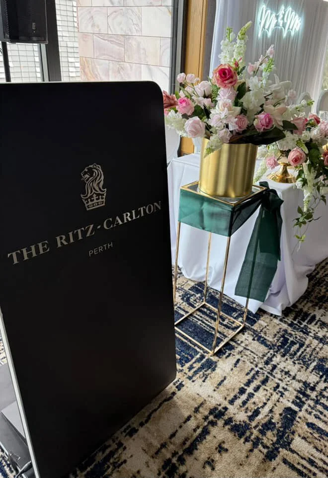 A black standing sign with the logo and text for The Ritz-Carlton Perth, positioned near a table with a gold vase and a large bouquet of pink and white flowers, in a decorated indoor setting.