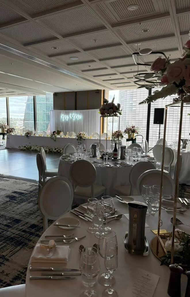 Elegant wedding reception setup in a high-rise venue with large windows, decorated with white and blush floral arrangements, round tables with white tablecloths, and a neon sign in the background that reads "Mr & Mrs."