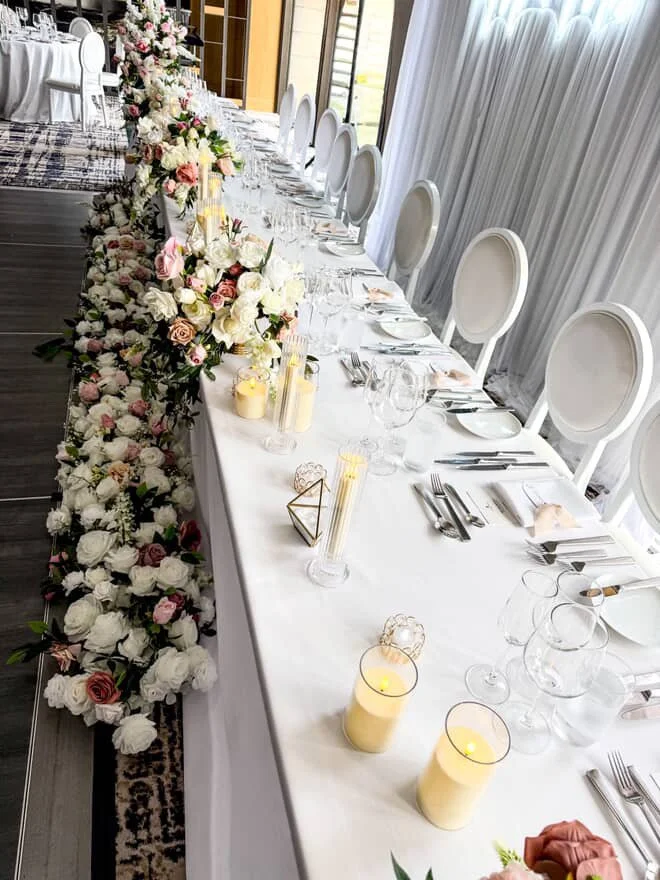 Long banquet table decorated with pink and white floral arrangements, candles, elegant tableware, and white chairs, set for a formal event in a bright room with white curtains.