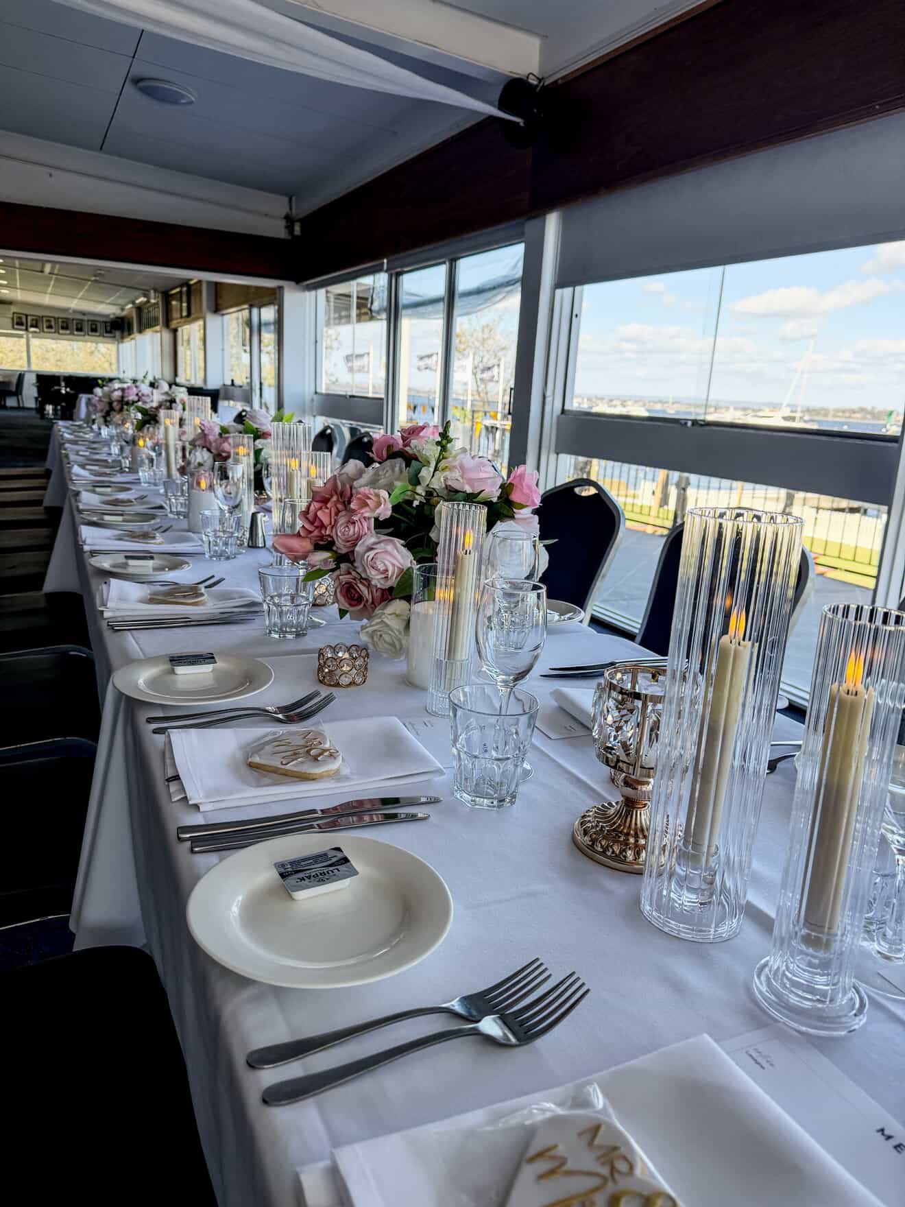 Elegant banquet table setup with white tablecloths, pink floral centerpieces, tall candles, and glassware, located in a room with large windows overlooking a sunny outdoor view.