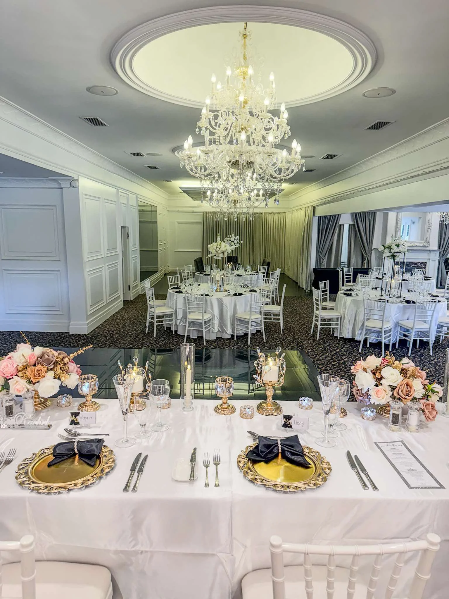 Elegant banquet hall decorated for a formal event with round tables covered in white tablecloths, floral centerpieces, and ornate gold and black place settings, with a large crystal chandelier hanging from the ceiling.