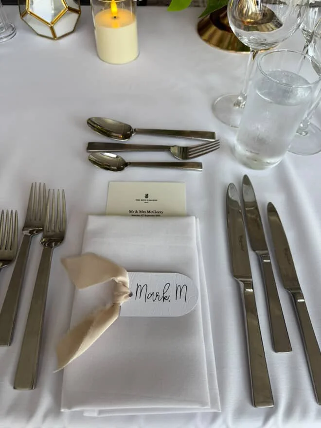 Formal dining table setup with multiple forks, knives, and spoons, a white napkin with a name card that reads "Mark M," and a white tablecloth. There are glasses of water, candles, and decorative items on the table.