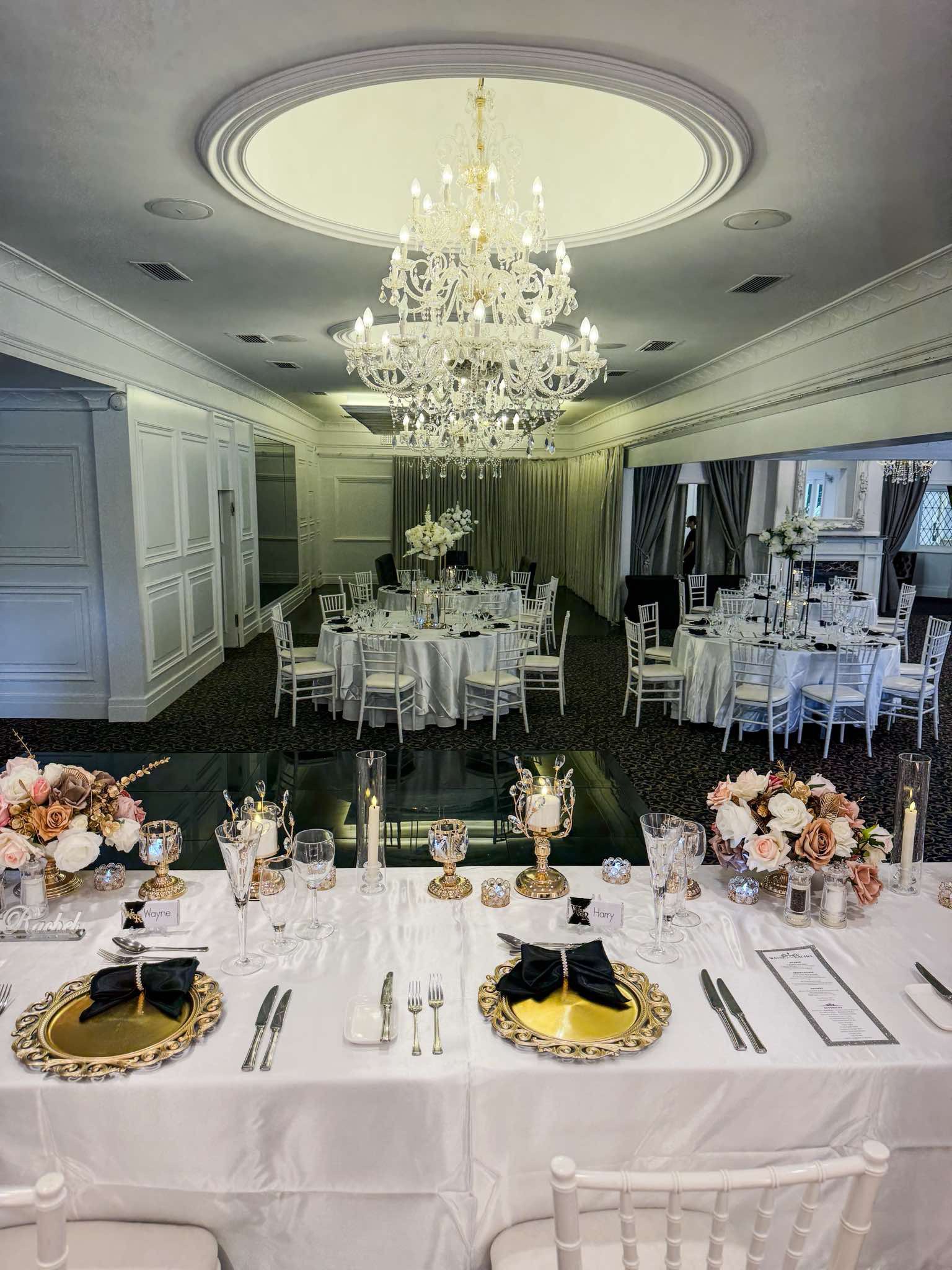 Elegant banquet table setup with gold-rimmed plates, black napkins, floral centerpieces, and a menu on a white tablecloth, with a formal event space in the background.