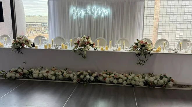 Long banquet table decorated with floral arrangements and candles, backdrop with white curtains, neon sign reading 'Menu,' windows showing outdoor scenery.