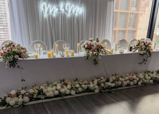 Long banquet table decorated with floral arrangements, candles, and white chairs at a wedding reception. There is a ceiling neon sign that reads 'Mr & Mrs' and a window showing a building outside.