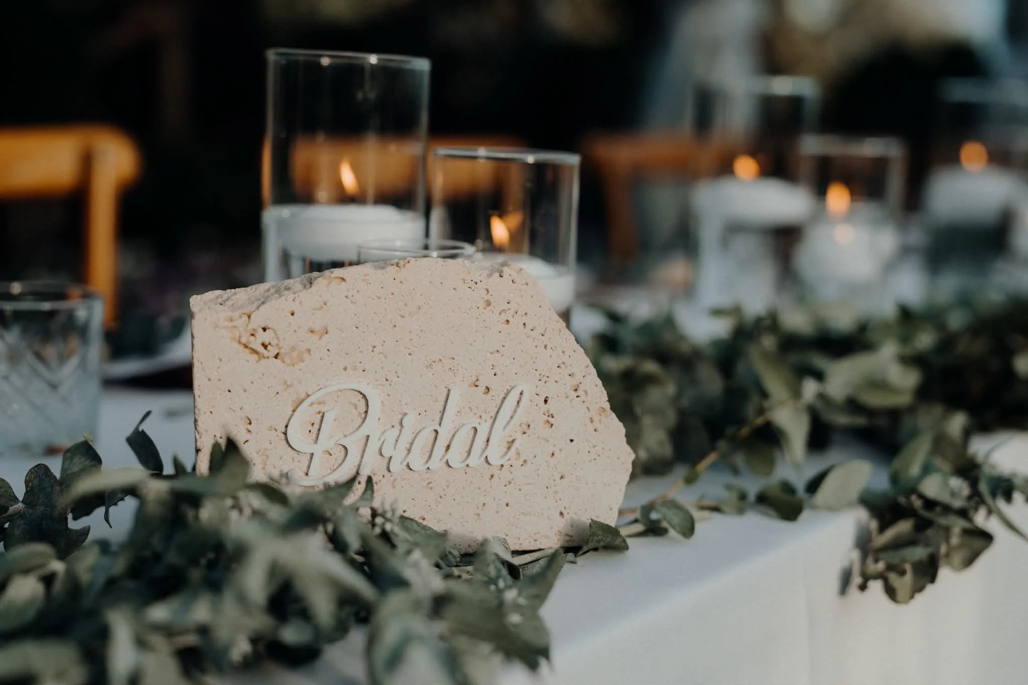 Decorative table setting with a beige stone with the word 'Bride' engraved on it, surrounded by green foliage, glassware, and candles in a dimly lit environment.