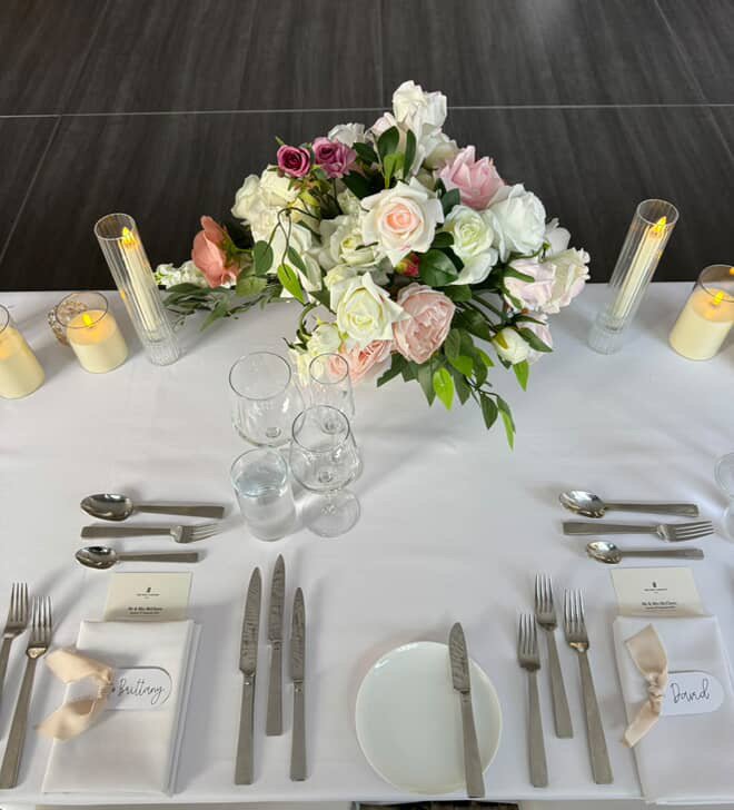 Table setting for a formal event with a large floral centerpiece of pink and white roses, lit candles in tall glass holders, water glasses, and neatly arranged silverware and napkins.