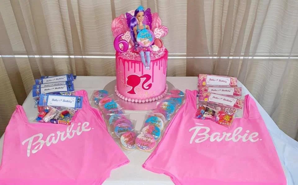 A pink birthday cake with a Barbie princess figurine on top, surrounded by pink and blue themed decorations, candies, and Barbie-branded pink bags on a white table.