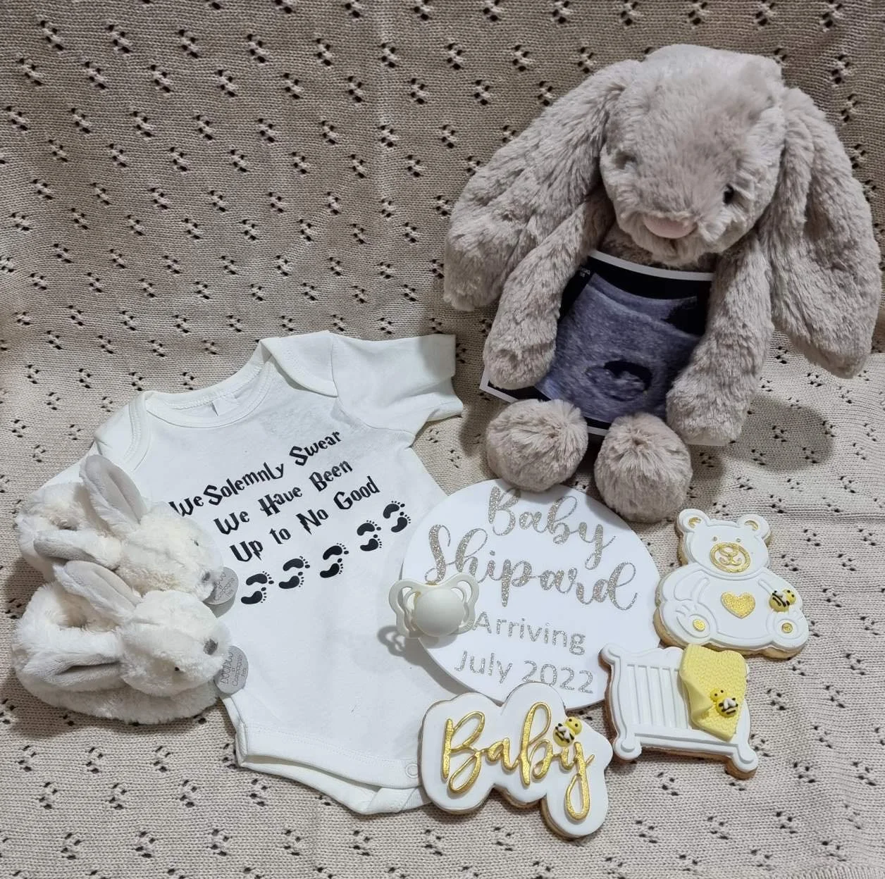 Baby shower gift set with plush bunny, baby clothes, decorated cookies, and baby-themed decorations on a textured beige surface.