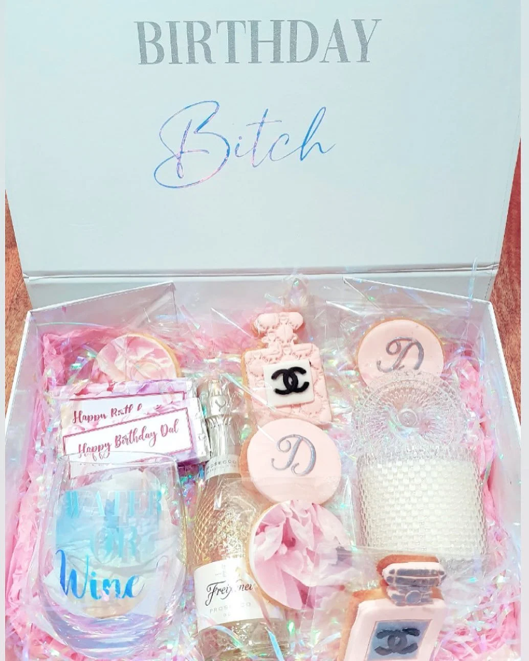 A birthday gift box decorated in pink with various themed items, including drinkware, cookies, and small decorative gifts, with a white interior lid displaying the words 'BIRTHDAY Bitch' in both uppercase and cursive handwriting.