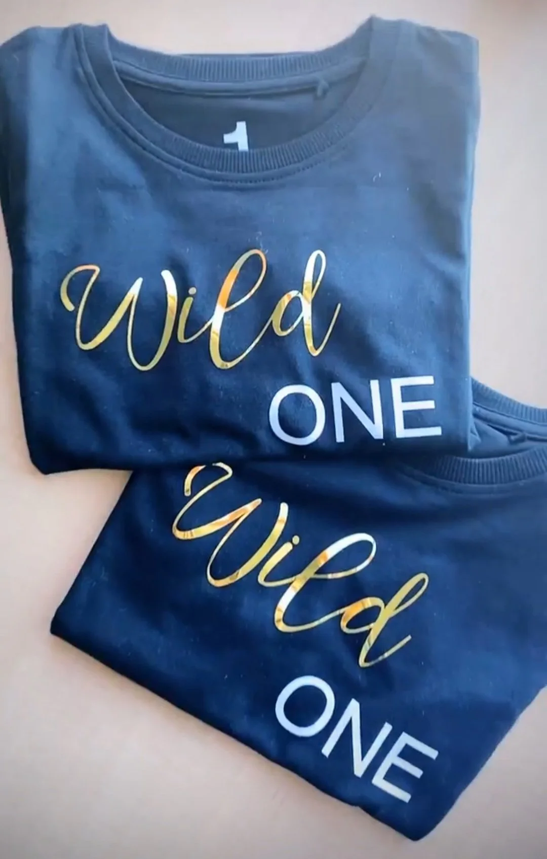 Two navy blue T-shirts with the words "Wild One" in gold and white lettering, laid flat. One shirt is overlapping the other.