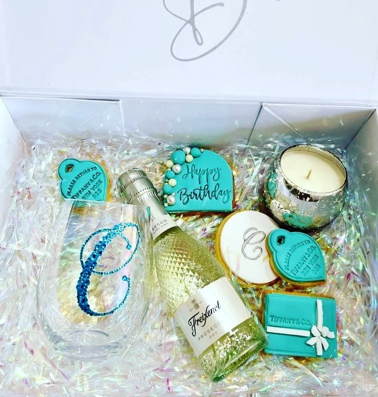A birthday gift box containing a Freixenet sparkling wine bottle, a clear wine glass with a blue letter, a teal candle holder with a candle, decorative teal and white cookies, a teal 2D and 3D party hat, and white shredded paper.