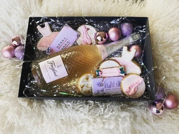 A gift box containing a bottle of champagne, Christmas cookies shaped like ornaments and other festive shapes, and pink and purple Christmas ornaments, all on a fluffy white surface.