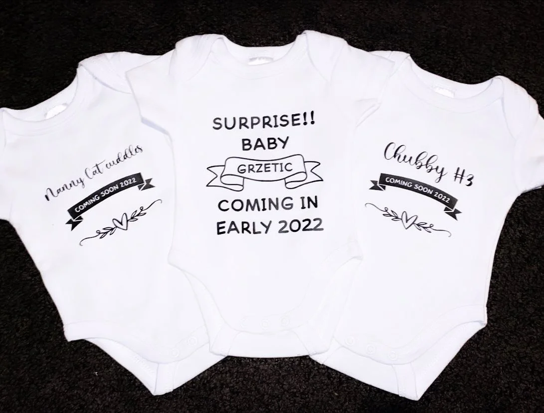 Three white baby onesies with black printed text and graphics on a black surface.