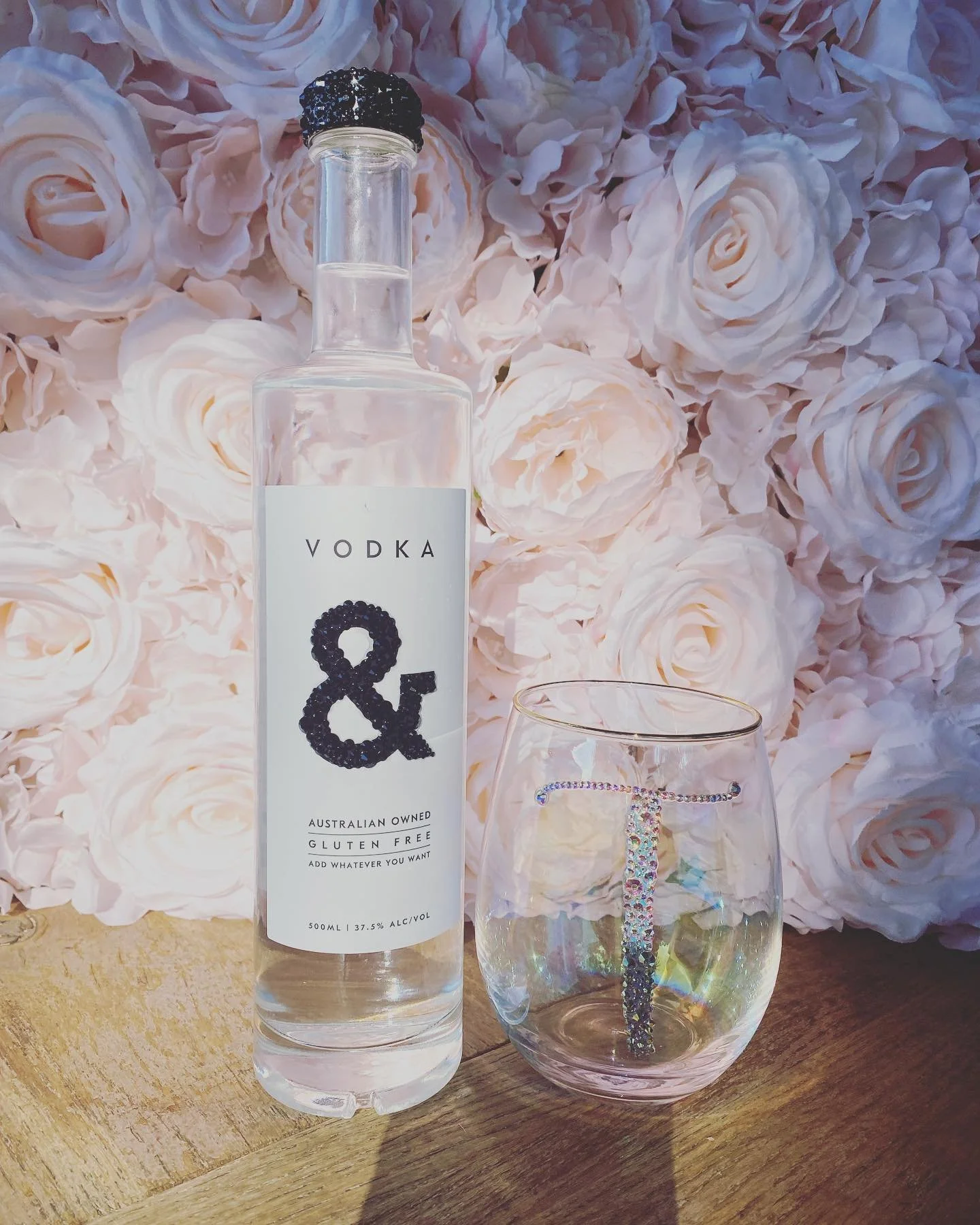 A bottle of vodka with a diamond-shaped ampersand logo on the label, a drinking glass with a crystal letter T inside, and a background of white roses.
