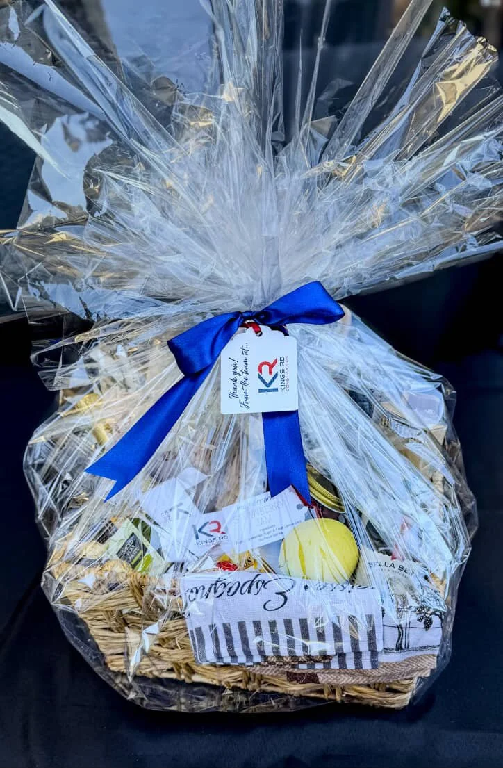 Gift basket wrapped in clear cellophane with a blue ribbon, containing assorted items and a thank you tag from Kings RD.