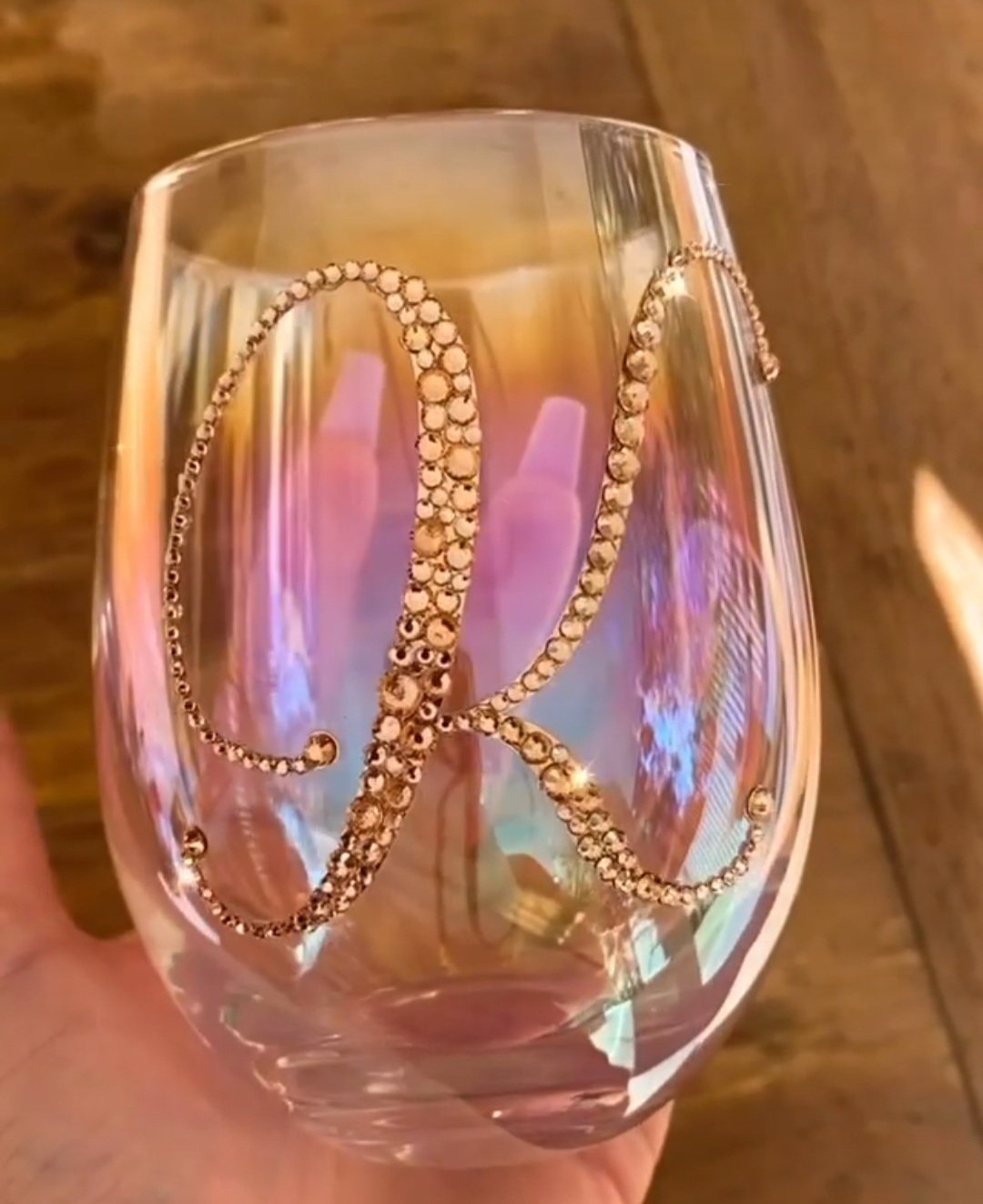 A clear glass with a gold cursive letter 'K' design made of rhinestones, held in someone's hand against a wooden background.