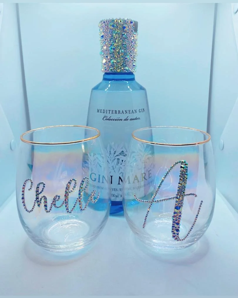 A bottle of Mediterranean gin with a sparkly, jeweled cap surrounded by two stemless glasses with gold rims. One glass has the word "Cheers" and the other has the word "Fun" written in rhinestones.