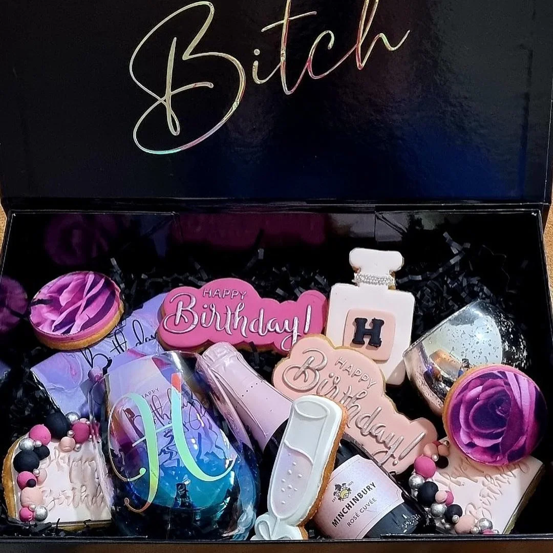 A birthday gift box containing decorated cookies with birthday themes, pink and purple roses-shaped cookies, a mini champagne bottle cookie, a thermometer-shaped cookie, a perfume-shaped cookie, and assorted pink, black, and silver beads.