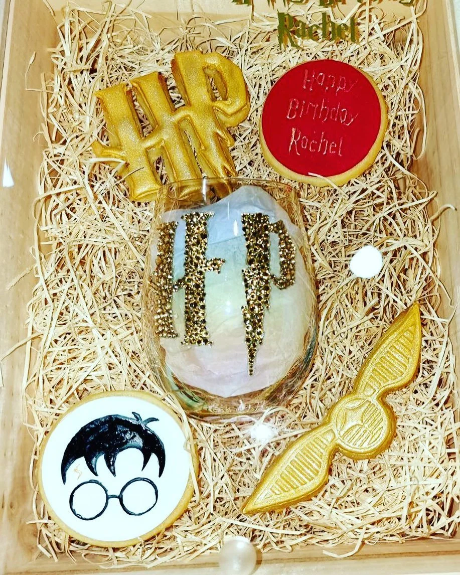 A birthday gift box with wood shavings containing decorated cookies and a glass of champagne. The cookies spell out 'HAPPY', 'TED', and have Harry Potter themed designs, including glasses, lightning bolt, and wings. There is also a round red cookie w