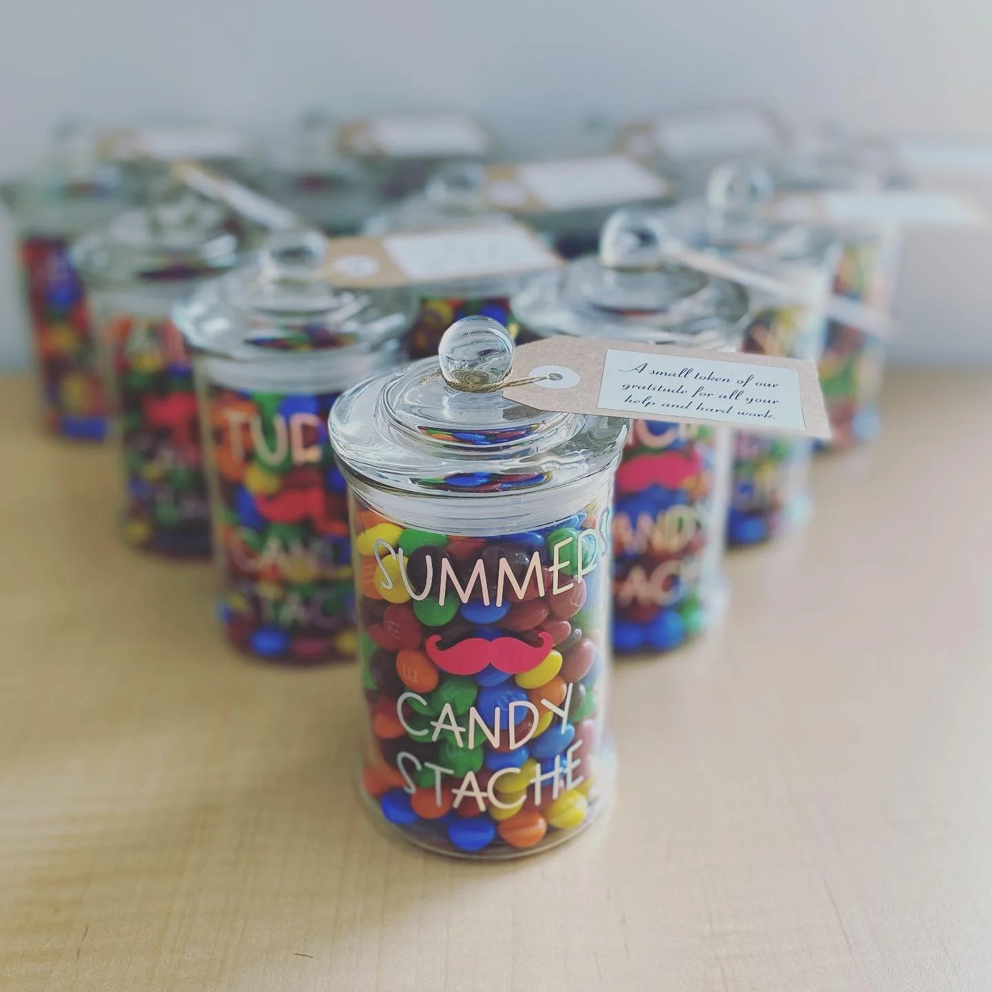 A jar filled with colorful candy with a label that reads 'Summer Candy Stache', and a small thank you tag attached that expresses gratitude for help and hard work, with multiple similar jars in the background.