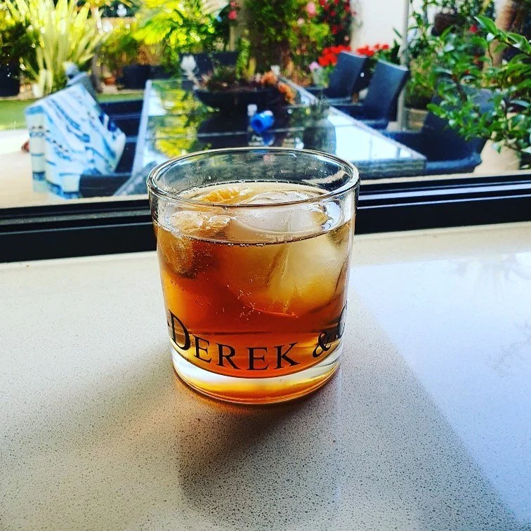 A glass of dark-colored beverage with ice cubes, placed on a windowsill with a garden and outdoor furniture visible outside.
