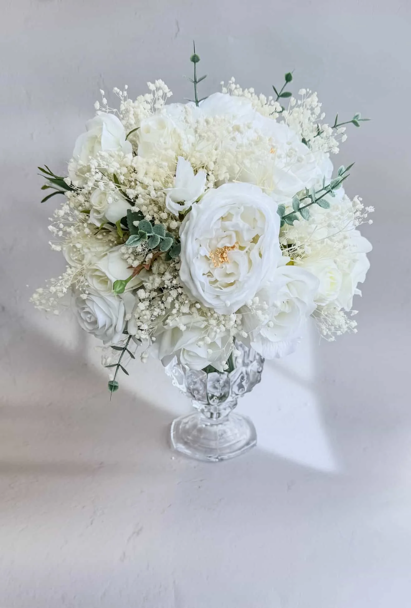 A bouquet of white flowers, including roses and peonies, arranged in a decorative glass vase against a plain background.