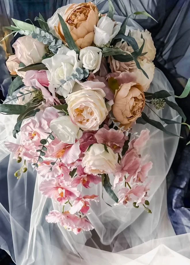 A bouquet of flowers including peonies, roses, and orchids with green leaves, wrapped in white tissue paper.