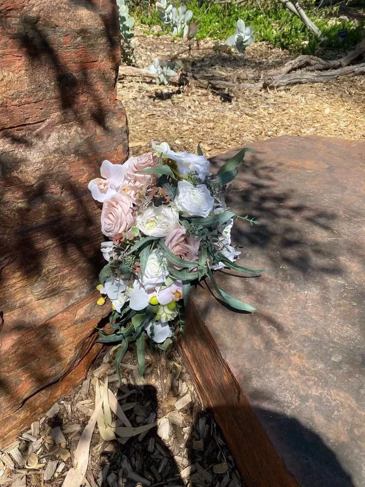 A bouquet of white and pale pink flowers resting on a large reddish-brown rock outdoors. The background includes dry soil, a cactus, and sunlight casting shadows.