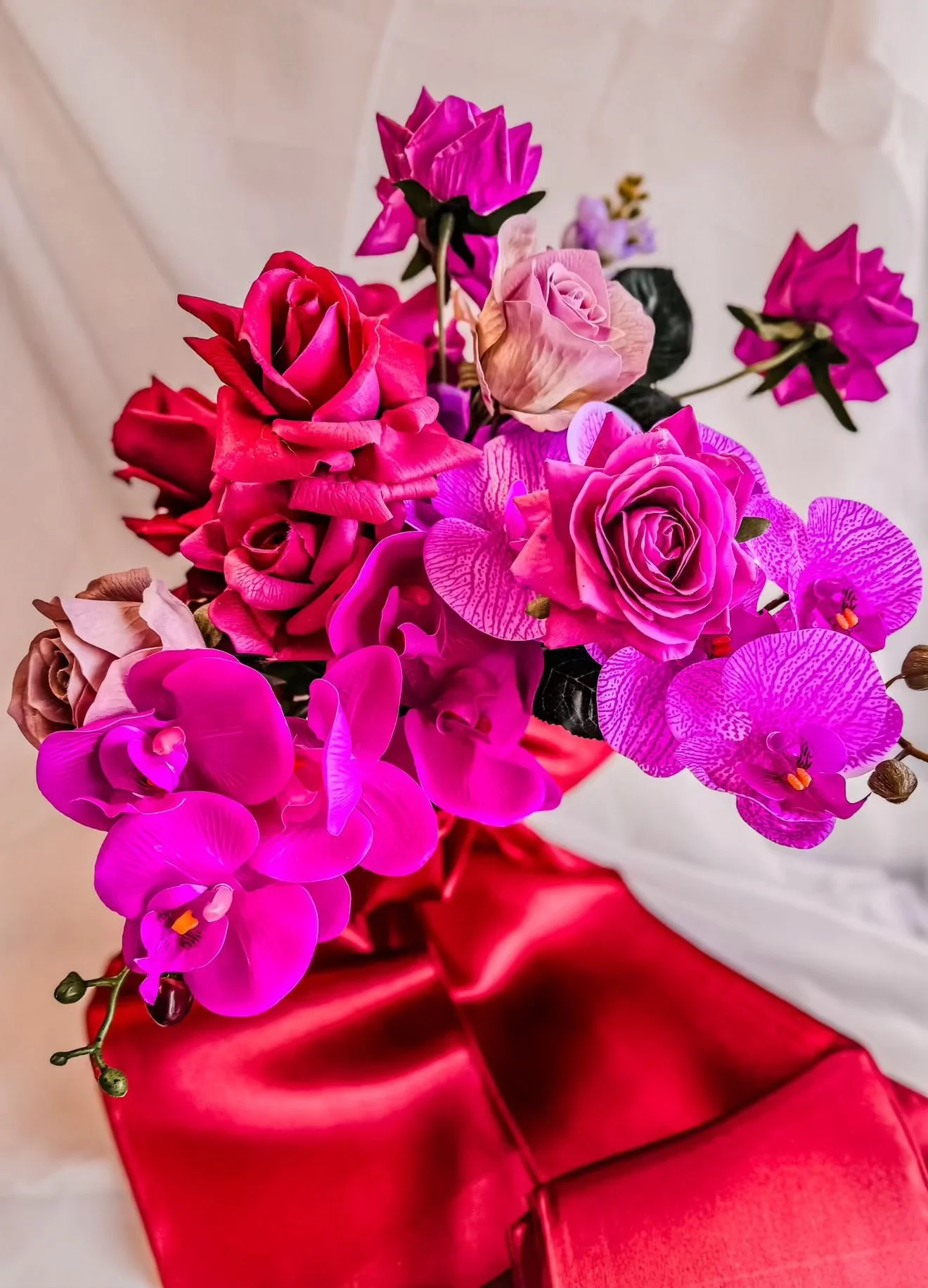 A bouquet of pink, purple, and peach flowers, including roses, orchids, and bougainvillea, wrapped in red paper, is displayed on a white surface.