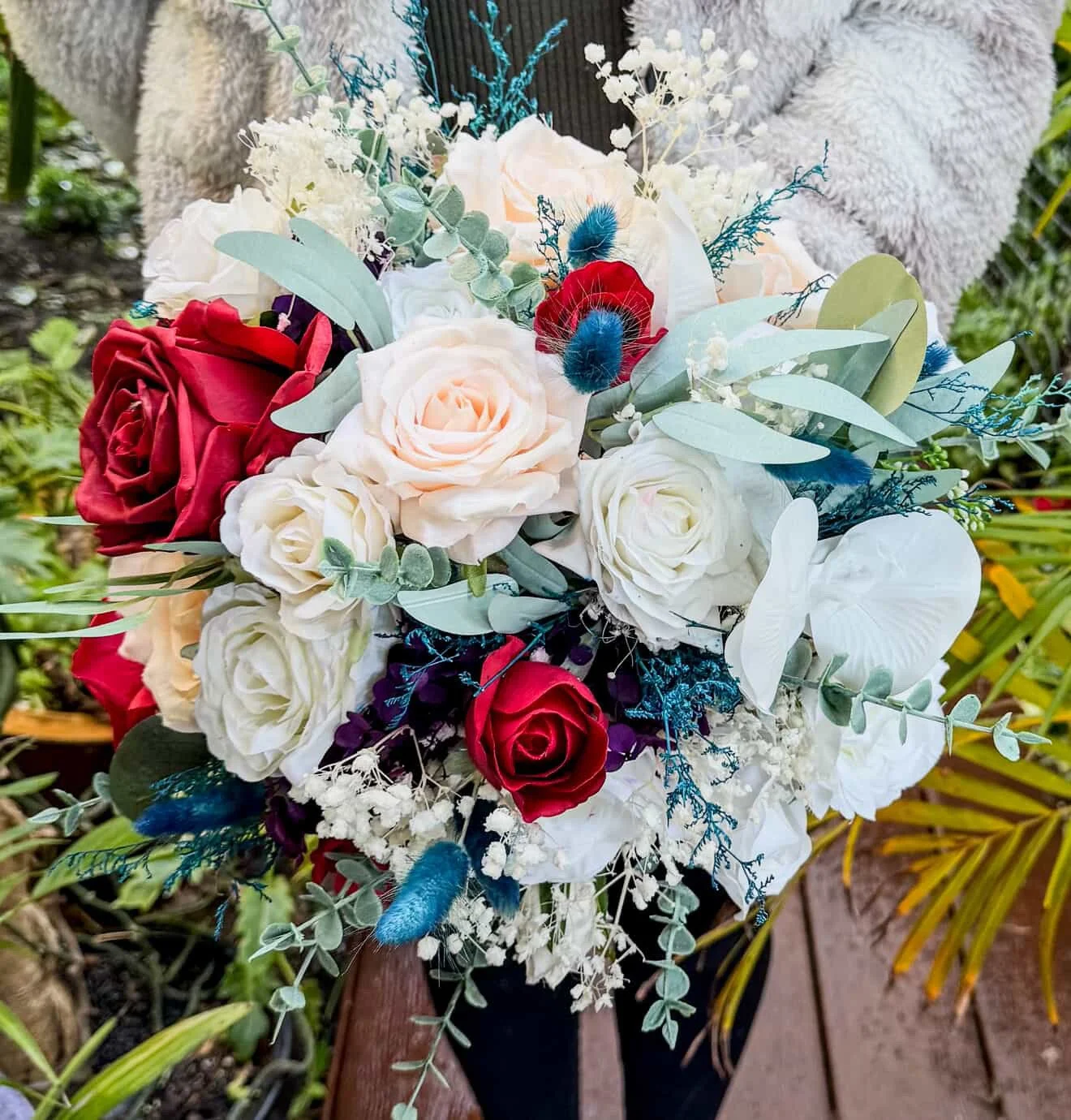 A person holding a bouquet of assorted flowers including red and white roses, white hydrangeas, and blue accents, with greenery and a blurred outdoor garden background.