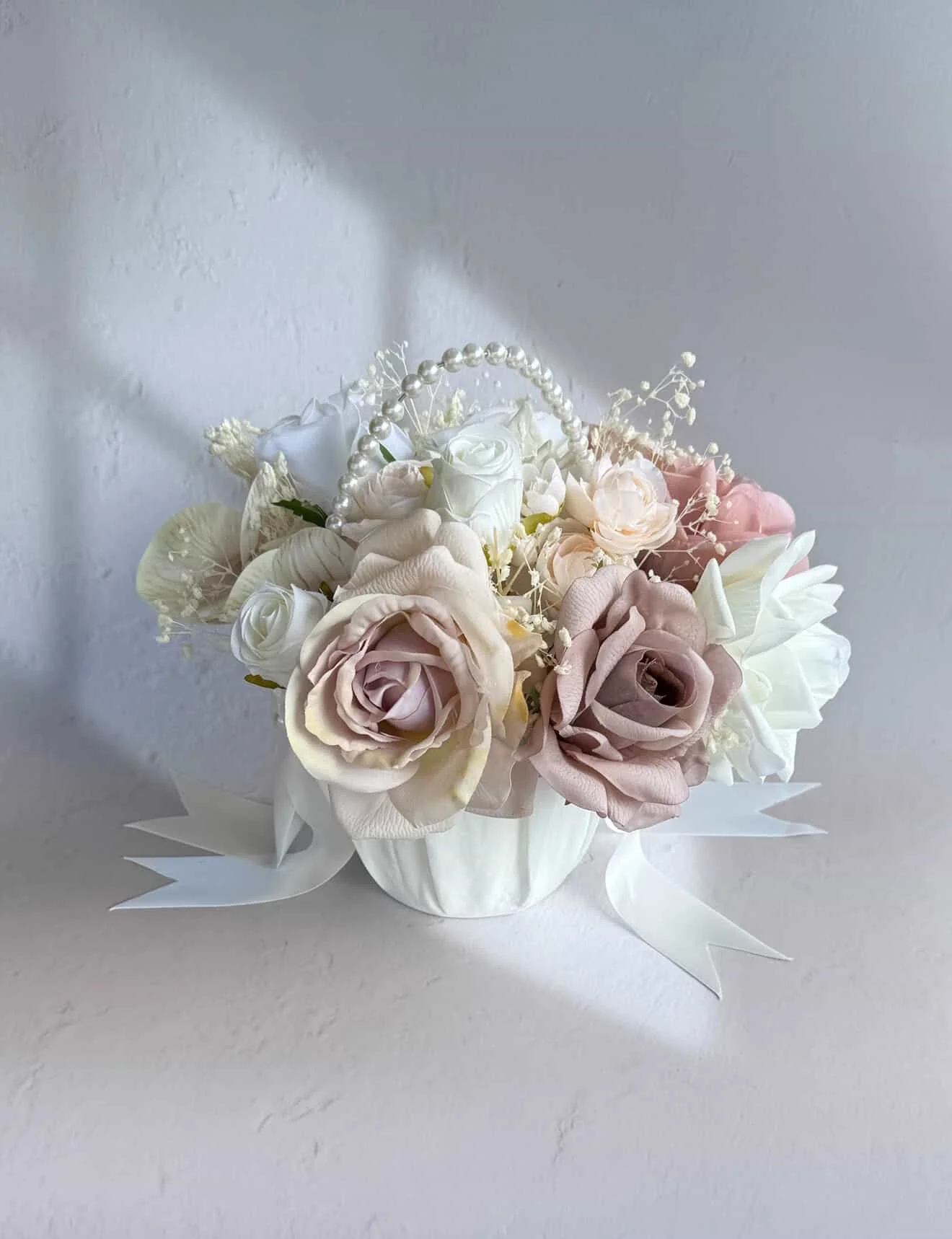 A floral arrangement with white and pastel pink roses, white lilies, and decorative white pearls in a white container, against a plain background.
